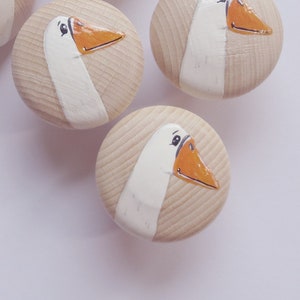 Könnte beinhalten: Fünf Holzknaufknöpfe mit bemalten Gänsegesichtern. Jeder Knopf zeigt eine weiße Gans mit orangefarbenem Schnabel und schwarzen Augen. Die Knöpfe sind hellbraun.