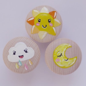 Könnte beinhalten: Drei Holzknöpfe mit bemalten Designs. Der erste Knopf zeigt eine gelbe Sonne mit einem lächelnden Gesicht. Der zweite Knopf zeigt eine weiße Wolke mit einem lächelnden Gesicht und Regentropfen. Der dritte Knopf zeigt einen gelben Halbmond mit einem schlafenden Gesicht.