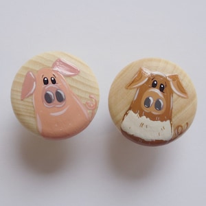 Könnte beinhalten: Zwei Holzknöpfe mit bemalten Schweinegesichtern. Das Schwein links ist rosa mit einer braunen Schnauze und das Schwein rechts ist braun mit einer weißen Schnauze.