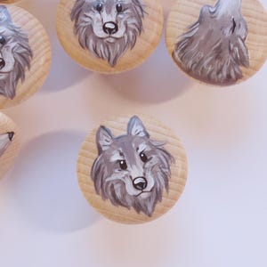 Könnte beinhalten: Eine Sammlung von Holzschrankknöpfen mit handgemalten Wolfsmotiven. Die Knöpfe sind rund mit einer natürlichen Holzoberfläche. Die Wölfe sind in Grau- und Weißtönen dargestellt. Einige heulen.