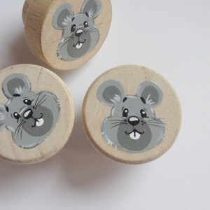 Könnte beinhalten: Drei Holzknöpfe mit bemalten grauen Mäusegesichtern. Die Knöpfe sind rund und haben eine hellbraune Farbe.
