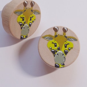 Könnte beinhalten: Zwei Holzknöpfe mit einem bemalten Giraffenkopf. Die Giraffe hat gelbes Fell mit schwarzen Flecken und eine braune Nase. Die Knöpfe sind rund und haben eine hellbraune Farbe.