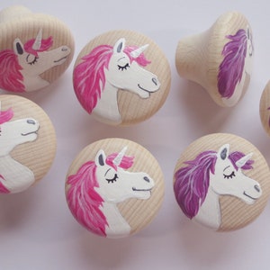 Könnte beinhalten: Acht Holzknauf für Schubladen, die mit Einhorn-Gesichtern bemalt sind. Die Einhörner haben rosa und lila Mähnen und weiße Gesichter mit geschlossenen Augen.
