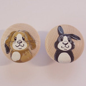 Könnte beinhalten: Zwei Holzknöpfe mit bemalten Tiergesichtern. Der erste Knopf zeigt einen braunen Hund mit schwarzen Ohren und einer weißen Brust. Der zweite Knopf zeigt ein schwarzes Kaninchen mit weißen Ohren und einer weißen Brust.