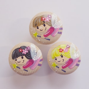 Könnte beinhalten: Drei runde Holzornamente mit bemalten Feendesigns. Jede Fee hat rosa Flügel, ein rosa Kleid und einen Zauberstab. Die Feen haben verschiedene Haarfarben: braun, blond und schwarz.