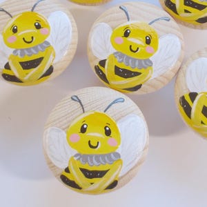 Könnte beinhalten: Sechs runde Holzschrankknöpfe mit handgemalten Bienenmotiven. Jeder Knopf zeigt eine gelbe Biene mit schwarzen Streifen, weißen Flügeln und einem lächelnden Gesicht. Die Knöpfe sind vor einem hellen Hintergrund angeordnet.