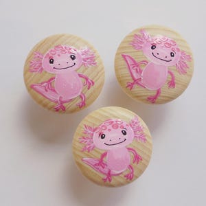 Könnte beinhalten: Drei hölzerne Schrankknöpfe mit hellen Holzmaserungen. Jeder Knopf zeigt eine rosa Axolotl-Illustration. Die Axolotl haben rosa Kiemen, Gliedmaßen und ein lächelndes Gesicht. Die Knöpfe sind rund und haben eine glatte Oberfläche.