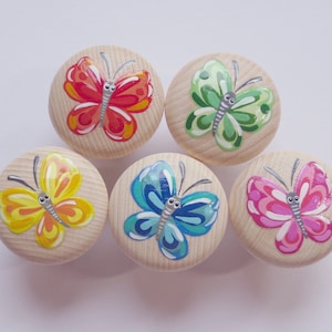 Könnte beinhalten: Fünf Holzknöpfe mit bemalten Schmetterlingsmotiven. Die Schmetterlinge sind in verschiedenen Farben: rot, grün, gelb, blau und pink.
