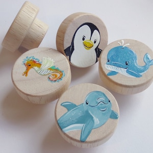 Könnte beinhalten: Fünf Holzgriffe mit bemalten Meeresbewohnern. Die Griffe zeigen einen Pinguin, einen Wal, ein Seepferdchen und zwei Delfine. Die Griffe sind in leuchtenden Farben bemalt und wären perfekt für ein Kinderzimmer.