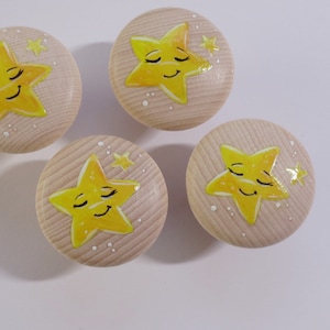 Könnte beinhalten: Vier Holzschrankknöpfe mit heller Holzmaserung. Jeder Knopf zeigt einen gelben Stern mit einem lächelnden Gesicht und kleinen weißen Punkten. Die Sterne haben geschlossene Augen, was auf ein schläfriges oder skurriles Thema hindeutet.