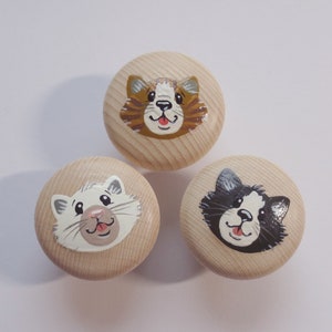 Könnte beinhalten: Drei Holzknöpfe mit bemalten Katzenköpfen. Die Katzenköpfe sind weiß, braun und schwarz mit weißen Akzenten. Die Knöpfe sind rund und haben eine helle Holzoptik.