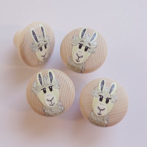 Könnte beinhalten: Vier hölzerne Schrankknöpfe mit handgemalten Lama-Gesichtern. Die Knöpfe sind hellbraun mit einem kreisförmigen Design, das ein Lama mit grauem Fell, schwarzen Augen und einem hellgrünen Gesicht zeigt. Ein Knopf hat eine Standardform.