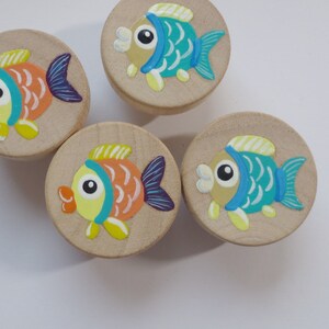 Könnte beinhalten: Vier Holzknöpfe mit bemalten Fischmotiven. Jeder Knopf zeigt einen Fisch in einer anderen Farbe mit gelben, orangefarbenen, blauen und grünen Details.
