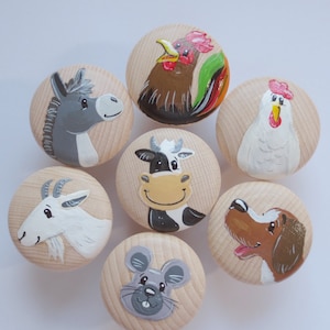 Könnte beinhalten: Satz von sieben Holzgriffen, die mit verschiedenen Bauernhoftieren bemalt sind. Die Tiere umfassen einen Hahn, eine Kuh, einen Esel, eine Ziege, ein Huhn, einen Hund und eine Maus. Die Griffe sind alle rund und haben eine glatte, natürliche Holzoptik.