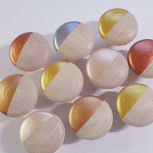 Könnte beinhalten: Eine Sammlung von zehn Holzschrankknöpfen. Jeder Knopf ist rund und hat auf einer Hälfte eine natürliche Holzmaserung und auf der anderen Hälfte eine metallische Lackierung in Gold-, Kupfer-, Silber- und Roségoldtönen. Die Knöpfe liegen auf einer weißen Oberfläche.