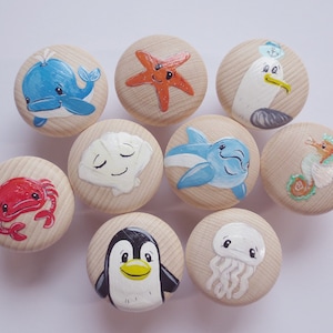 Könnte beinhalten: Zehn hölzerne Schrankknöpfe, jeder handbemalt mit einem anderen Meereskreaturen-Design. Die Knöpfe zeigen einen Wal, einen Seestern, eine Möwe, eine Krabbe, eine Muschel, einen Delfin, ein Seepferdchen, einen Pinguin und eine Qualle. Die Knöpfe sind rund.