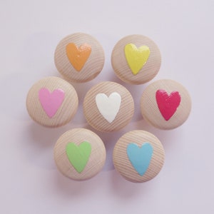 Könnte beinhalten: Sieben Holzknöpfe mit bemalten Herzen in verschiedenen Farben: orange, gelb, rosa, weiß, rot, grün und blau.