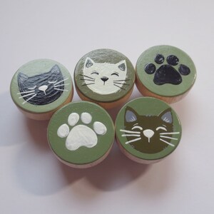 Könnte beinhalten: Fünf grüne Holz Kreise mit gemalten Katzen- und Pfotenabdrücken. Die Kreise sind in einem gedeckten Grünton bemalt. Die Katzenabdrücke sind schwarz, weiß und braun. Die Pfotenabdrücke sind schwarz und weiß.