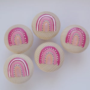 Könnte beinhalten: Fünf Holzknaufknöpfe mit bemalten Regenbogenmustern. Jeder Knopf zeigt einen rosa, orangefarbenen und weißen Regenbogen mit weißen Punkten.