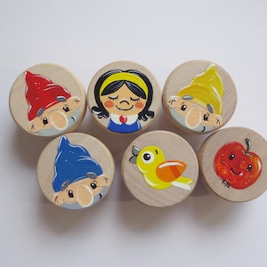 Könnte beinhalten: Sechs Holzrundungen mit bemalten Bildern. Die Bilder zeigen ein Mädchen mit dunklen Haaren, einen Zwerg mit roter Mütze, einen Zwerg mit gelber Mütze, einen Zwerg mit blauer Mütze, einen gelben Vogel und einen roten Apfel.