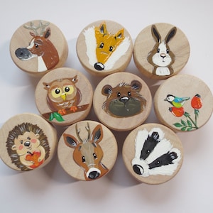 Könnte beinhalten: Satz von neun runden Holz-Ornamenten, die mit verschiedenen Waldtieren bemalt sind, darunter ein Reh, ein Fuchs, ein Kaninchen, eine Eule, ein Bär, ein Vogel, ein Igel und ein Dachs. Die Ornamente sind in leuchtenden Farben bemalt und haben einen verspielten Stil.