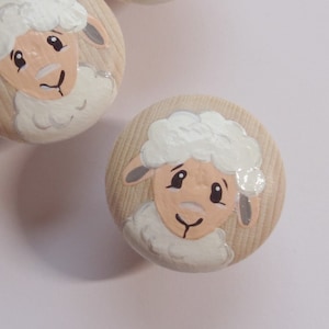 Könnte beinhalten: Drei Holzknöpfe mit bemalten Schafgesichtern. Jeder Knopf zeigt ein weißes Schaf mit einem hellbraunen Gesicht und schwarzen Augen. Die Schafe sind auf einem hellbraunen Holzhintergrund bemalt.
