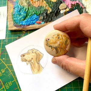 Könnte beinhalten: Ein Holzrund mit einer gemalten Illustration eines Hundekopfes, gehalten von einer Hand. Der Hund ist hellbraun mit einer rosa Nase. Ein Bleistift und eine Skizze des Hundes sind ebenfalls sichtbar.