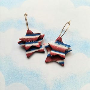 May include: A pair of star-shaped earrings with red, white, and blue stripes. The earrings are made of a layered material and hang from gold-colored hoops. The background is a light blue and white watercolor design.