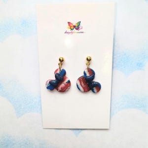 May include: A pair of handmade earrings with a red, white, and blue marbled design. The earrings have a Mickey Mouse-inspired shape with a gold-colored stud and a glittery blue accent. The earrings are displayed on a white card with a colorful butterfly logo.