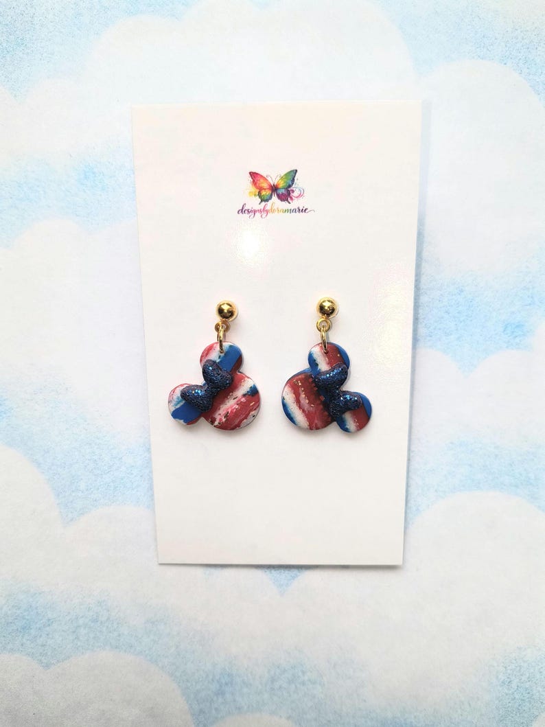 May include: A pair of handmade earrings with a red, white, and blue marbled design. The earrings have a Mickey Mouse-inspired shape with a gold-colored stud and a glittery blue accent. The earrings are displayed on a white card with a colorful butterfly logo.