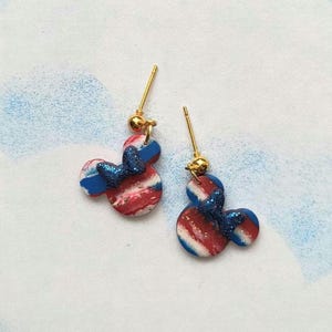 May include: A pair of stud earrings featuring a Mickey Mouse silhouette design. The earrings are red, white, and blue with a glittery blue bow. The posts and ball accents are gold.