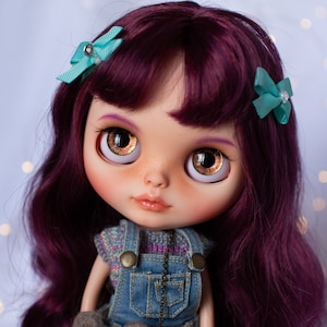May include: A doll with long, dark purple hair styled in loose waves. She is wearing a blue denim overall dress with a grey and white striped sweater underneath. She has brown eyes with gold flecks and is wearing two turquoise bows in her hair.