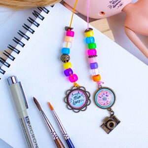 May include: A colorful beaded bookmark with two charms. One charm features a cat with the text "Min drumu" and the other charm features a donut with a camera hanging from it.