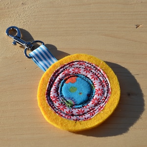 May include: A yellow felt circle with a multi-colored fabric design in the center. The fabric design features a blue circle with red and white flowers. The circle is attached to a silver key chain.