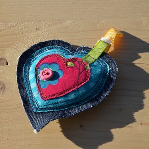 Puede incluir: Un adorno de tela en forma de corazón con capas de tela azul, rosa y roja. El adorno tiene un botón rosa con un diseño de flor y una cinta amarilla y verde.