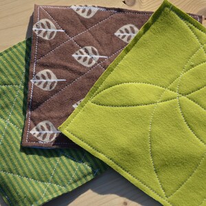 May include: Three quilted pot holders in green and brown with leaf patterns. The pot holders are made of fabric and have a quilted design.