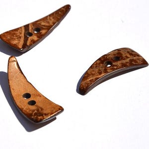 May include: Three brown, crescent-shaped buttons with two holes each, made of a natural material. The buttons have a unique, textured appearance with varying shades of brown and a glossy finish. They are arranged on a white surface.