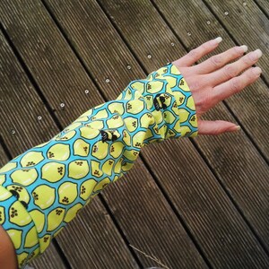 Pu&ograve; includere: Un manicotto a maniche lunghe con un motivo a limone. Il tessuto &egrave; giallo con un motivo a nido d'ape azzurro e illustrazioni di limoni neri. Il manicotto &egrave; indossato sul braccio.