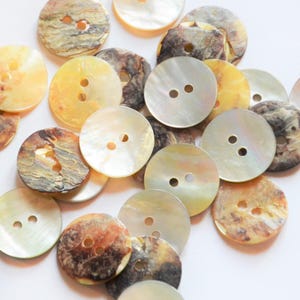 Puede incluir: Una colección de botones redondos en varios colores, incluyendo crema, amarillo y marrón. Los botones tienen dos agujeros para coser y un brillo natural e iridiscente. Los botones están hechos de un material natural.