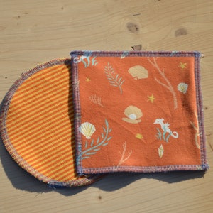 Puede incluir: Dos cuadrados de tela naranja y amarilla con ribete azul. Un cuadrado tiene un patrón a rayas y el otro tiene un patrón de caballitos de mar, conchas y algas marinas.