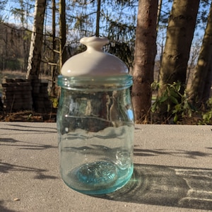 May include: A clear glass jar with a white ceramic lid. The jar has a light blue tint and is sitting on a grey surface.