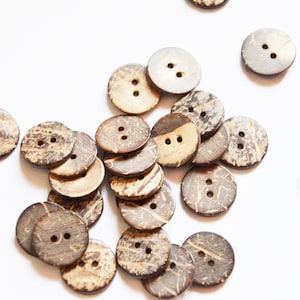 Könnte beinhalten: Eine Ansammlung runder Knöpfe aus natürlicher Kokosnussschale mit jeweils zwei Löchern. Die Knöpfe variieren in der Farbe von hellbraun bis dunkelbraun, mit sichtbaren Holzmaserungen. Sie liegen auf einer weißen Oberfläche verstreut.
