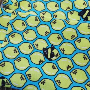 Pu&ograve; includere: Primo piano di tessuto con un motivo ripetuto di limoni gialli su sfondo blu brillante. Ogni limone ha dettagli di semi marroni. &Egrave; visibile anche un disegno in bianco e nero a forma di ape. Il tessuto &egrave; piegato.