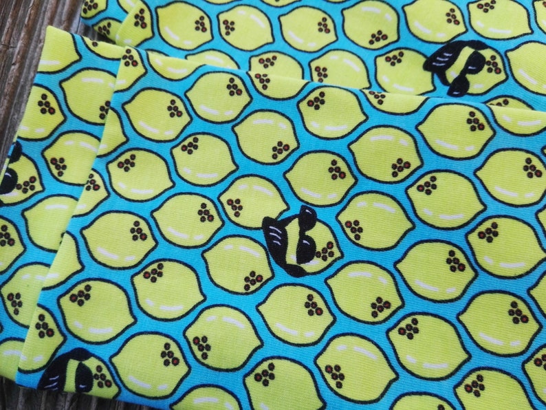 Pu&ograve; includere: Primo piano di tessuto con un motivo ripetuto di limoni gialli su sfondo blu brillante. Ogni limone ha dettagli di semi marroni. &Egrave; visibile anche un disegno in bianco e nero a forma di ape. Il tessuto &egrave; piegato.