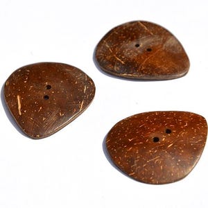 Puede incluir: Tres botones de coco marrones de forma irregular, cada uno con dos agujeros. Los botones tienen una superficie natural y texturizada, dispuestos sobre un fondo blanco. Estos botones son adecuados para proyectos de costura y artesanía.