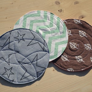 Può includere: Tre sottobicchieri rotondi in tessuto con motivi diversi. Un sottobicchiere ha uno sfondo blu con stelle nere, un altro ha un motivo a chevron verde e bianco, e l'ultimo ha uno sfondo marrone con stampe di foglie bianche.