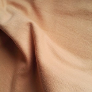 May include: A close-up of a soft, peach-colored fabric with a subtle texture. The fabric is folded and draped, creating a smooth, flowing look.
