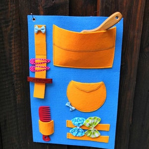 Peut inclure: Un tableau en feutre bleu avec différentes poches et boucles pour ranger des accessoires. Le tableau a un peigne, une cuillère en bois, une brosse à cheveux et plusieurs pinces et nœuds pour cheveux.