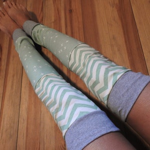 Op de afbeelding: Een paar groene en witte gestreepte beenwarmers met grijze manchetten. De beenwarmers hebben een chevron-patroon en een klein kruispatroon.