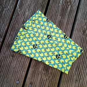 Pu&ograve; includere: Un oggetto di tessuto piegato con un motivo ripetuto di limoni gialli e api nere su sfondo azzurro. Il tessuto &egrave; appoggiato su una superficie di legno invecchiato.